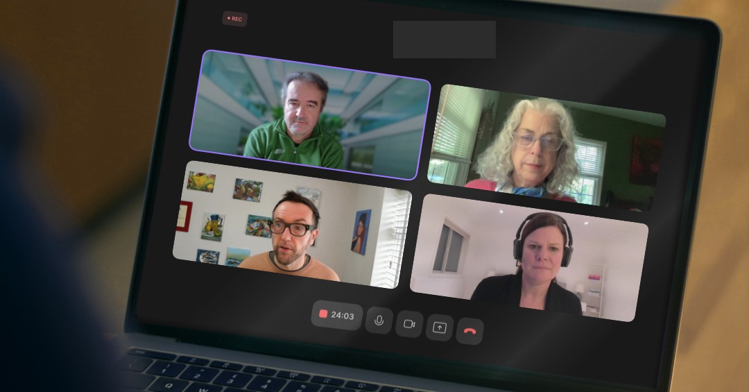 A laptop screen displays a video call with four people, each in their own window. The participants appear to be engaged in conversation, with various backgrounds behind them, including a green wall and some artwork.